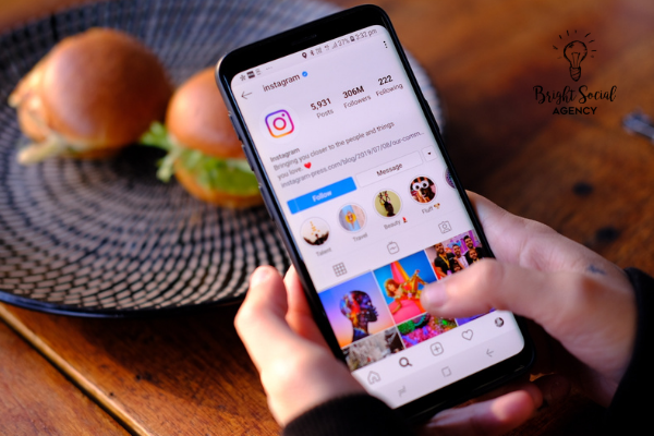 Hands holding a phone displaying the Instagram app. Two mini burgers on a plate in the background.
