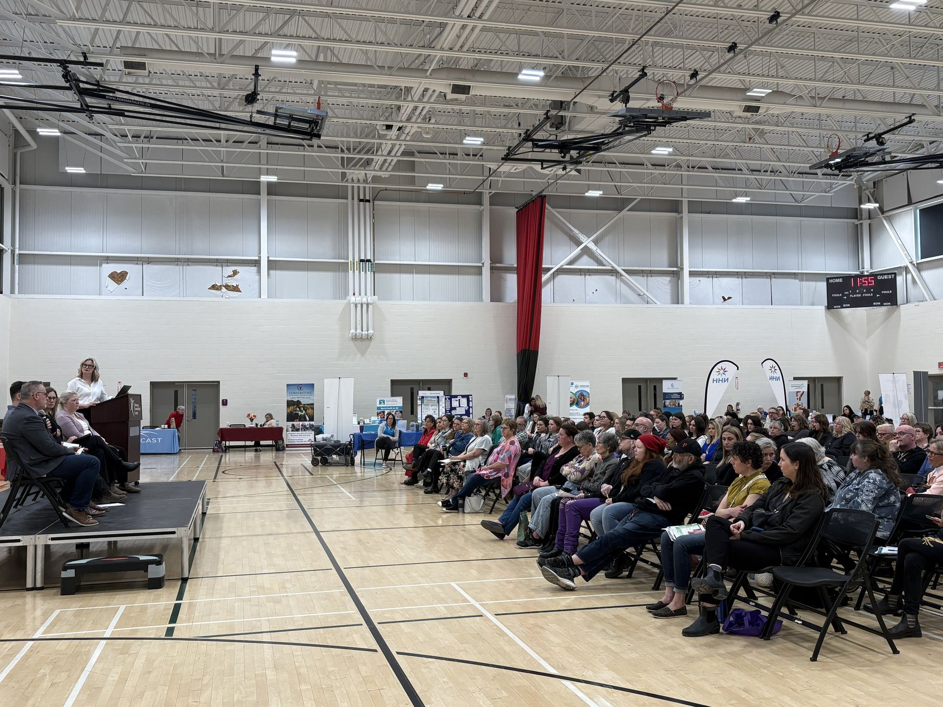 Audience seated in a gymnasium, watching speakers on a small stage.