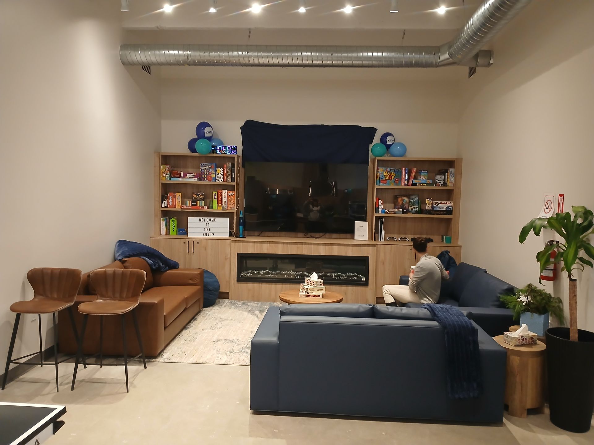 A lounge area with two sofas, two bar stools, shelves filled with items, a fireplace, and a person sitting on a sofa.
