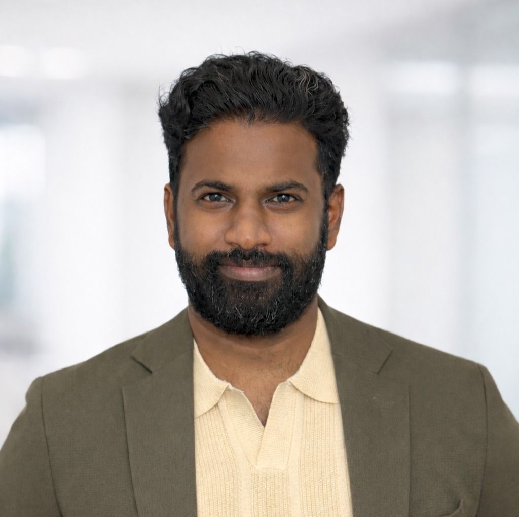 A person with a beard, wearing a light yellow polo shirt and an olive green blazer, stands against a blurry office background.