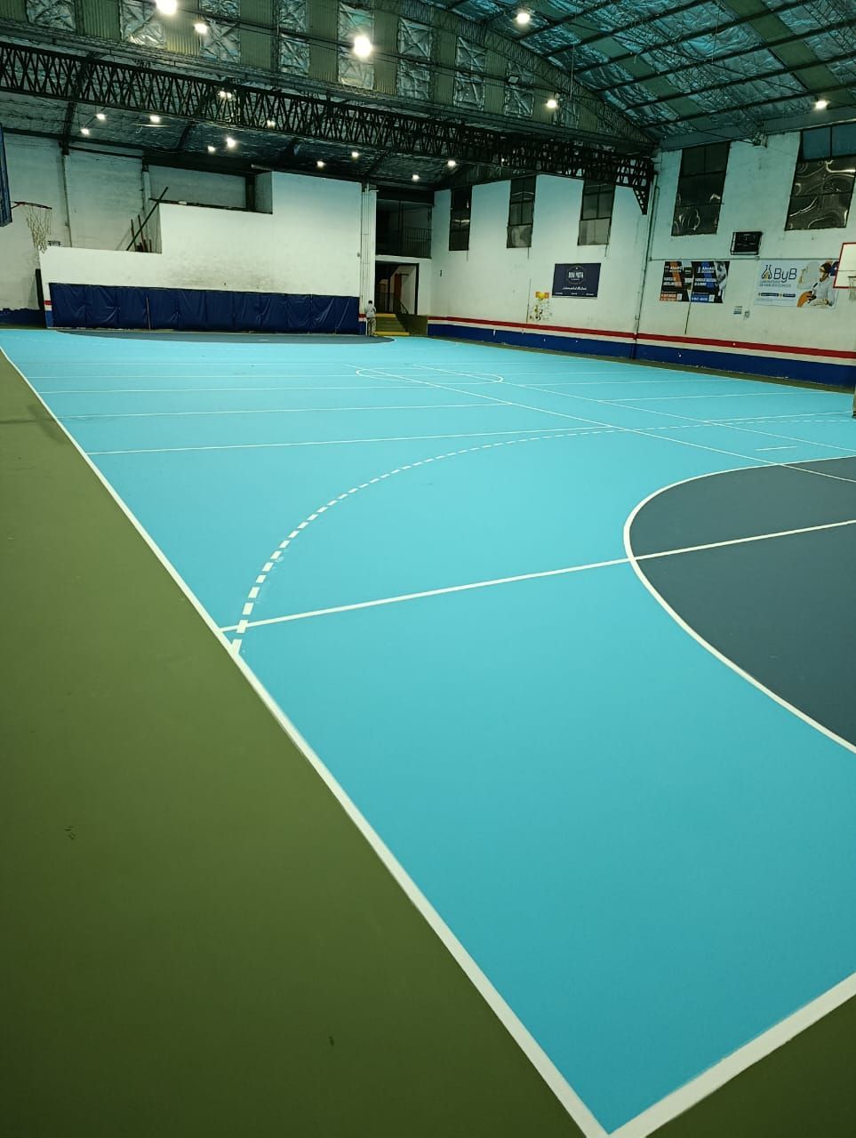 Pista deportiva cubierta vacía con suelo turquesa y verde bajo luces brillantes del techo.