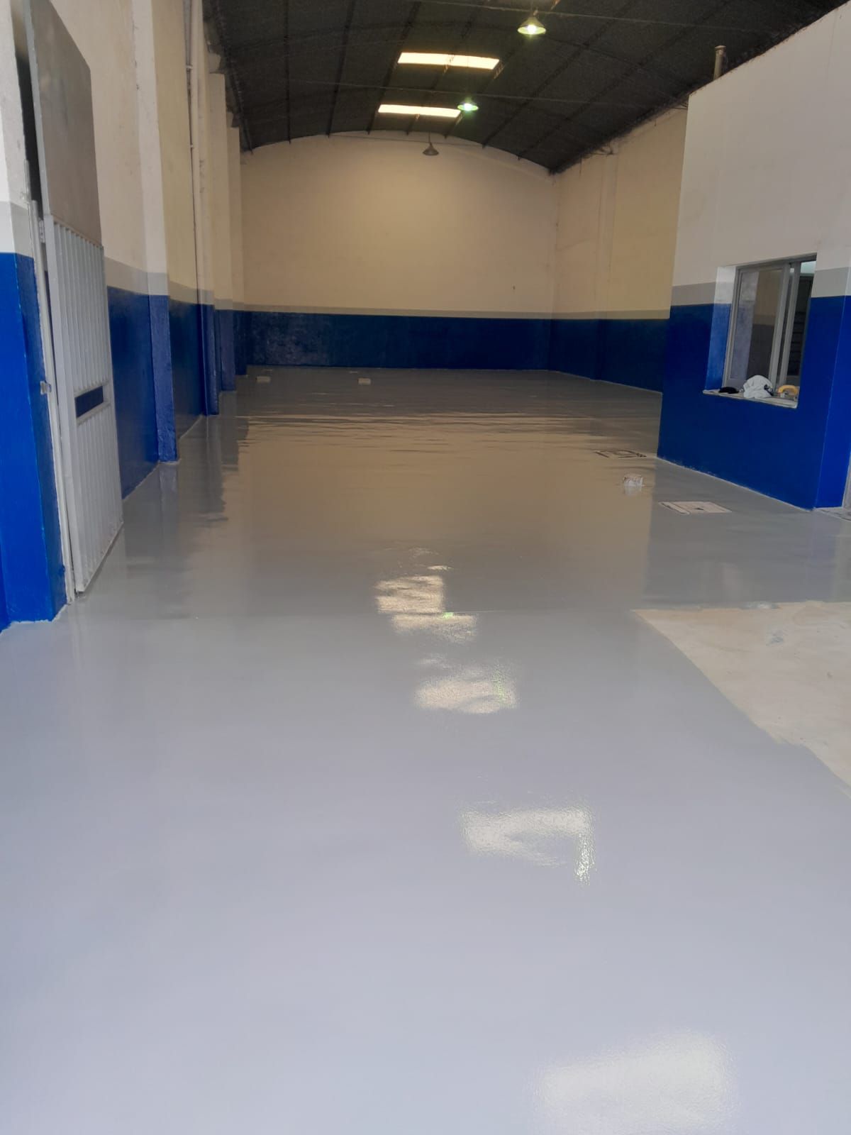Interior de un almacén vacío con suelo blanco brillante, paredes inferiores azules y luces en el techo.
