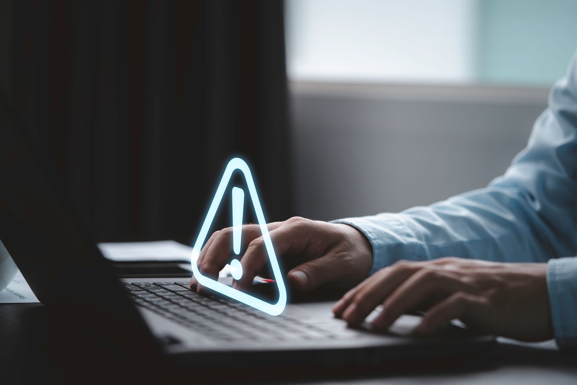 Hands typing on a laptop with a glowing warning icon on the screen in a dim room