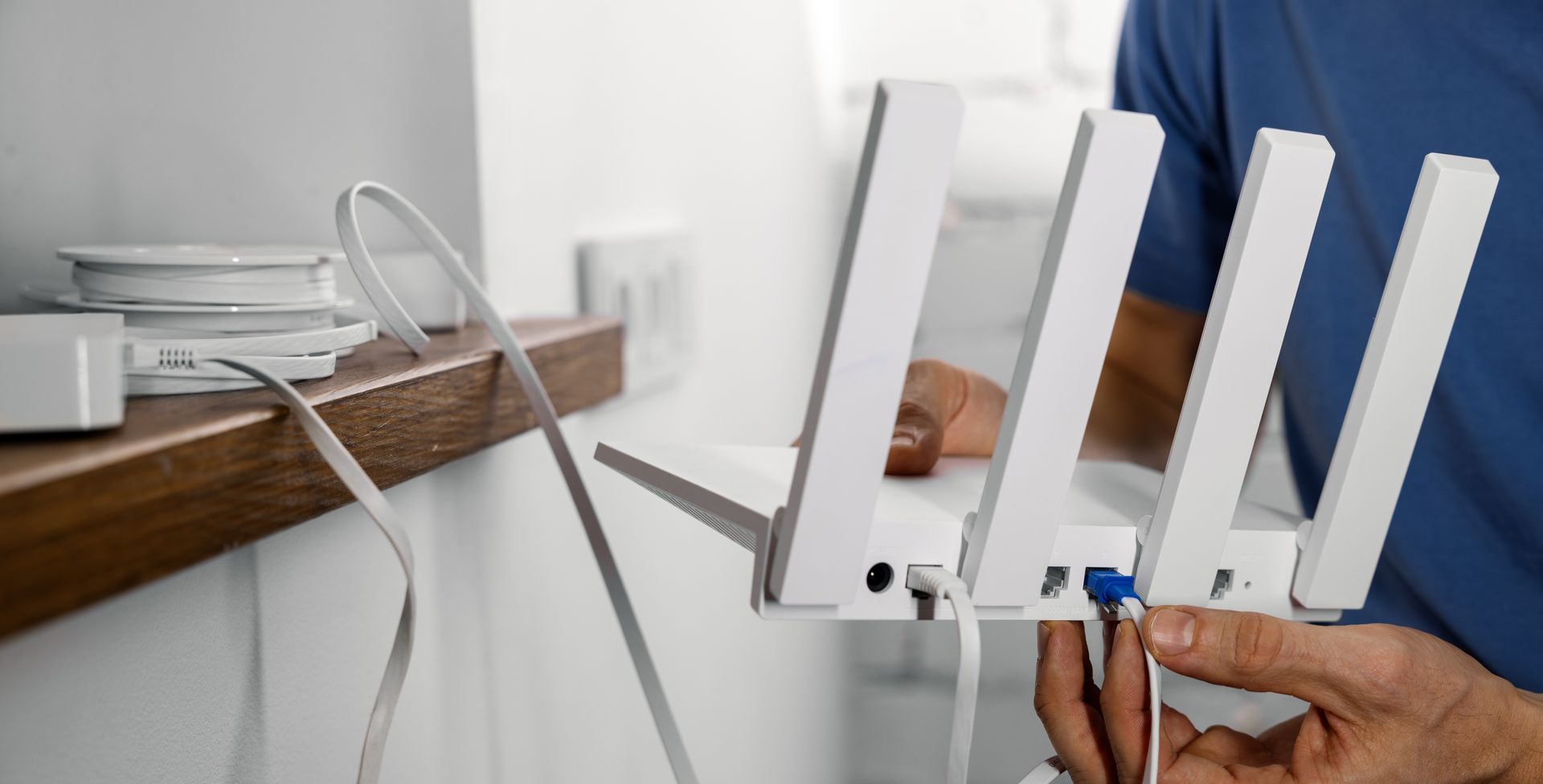 Person connects blue cable to a white Wi‑Fi router with antennas on a desk