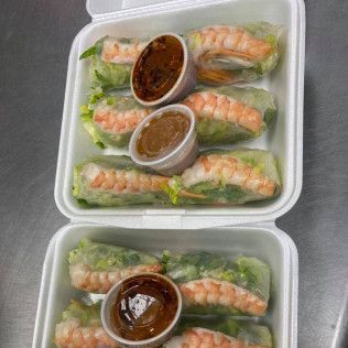 Two takeout containers of shrimp spring rolls with dipping sauces on a stainless steel counter