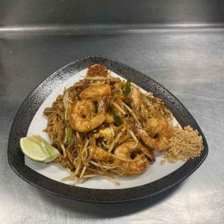Shrimp noodle stir-fry on a black plate with lime wedges and a side of chili paste