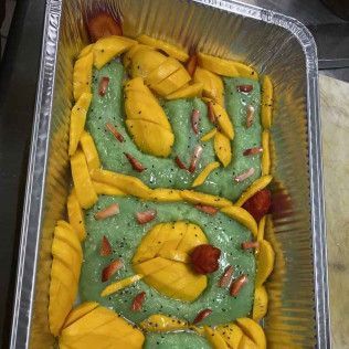 Foil tray of dessert topped with green and yellow swirls, mango slices, and red garnish