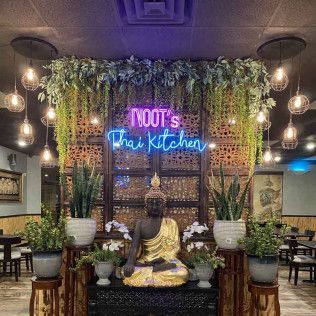 Thai restaurant interior with neon “NOOT’s Thai Kitchen” sign, hanging plants, and a seated Buddha statue