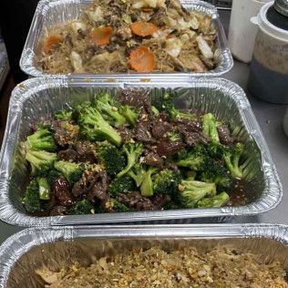 Three aluminum trays of cooked food, including broccoli beef, rice, and a vegetable dish.