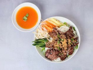 Bowl of rice with grilled beef, vegetables, and dipping sauce on a light table