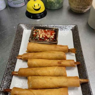 Five golden fried spring rolls on a square plate with dipping sauce in a restaurant setting