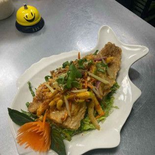 Grilled fish topped with herbs and shredded vegetables on a fish-shaped white plate, with a small bell nearby