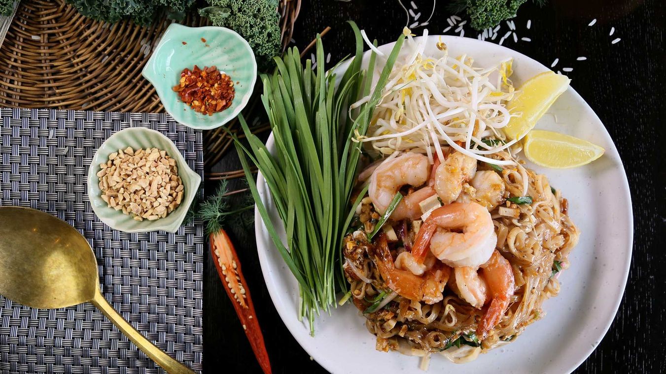 Thai noodle dish with shrimp, bean sprouts, herbs, lime, and chili on a white plate with dipping condiments