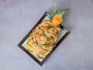 Grilled shrimp on rice in a rectangular black tray, garnished with pineapple leaves and an orange flower