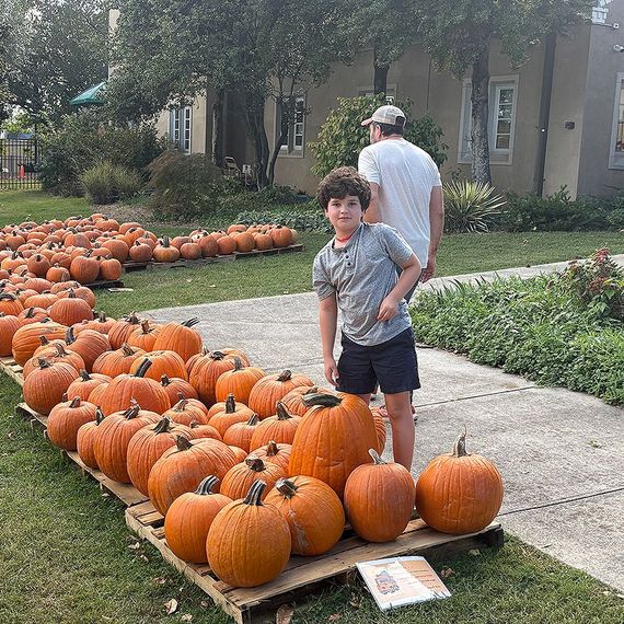 Boy in shorts and tee shirt smiles near pumpkins, adult in white shirt in background. Outdoors.