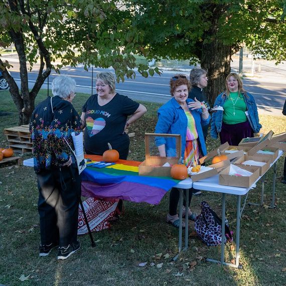 People at an outdoor LGBTQ+ event, pumpkins, rainbow flag. Some wearing pride shirts, some smiling.