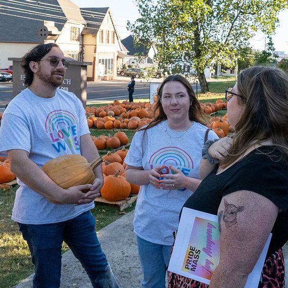 Three people with rainbow shirts at a pumpkin patch, smiling and talking. Houses are in the background.