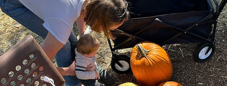 Woman and child looking at pumpkins near a black wagon, outdoors on a sunny day.