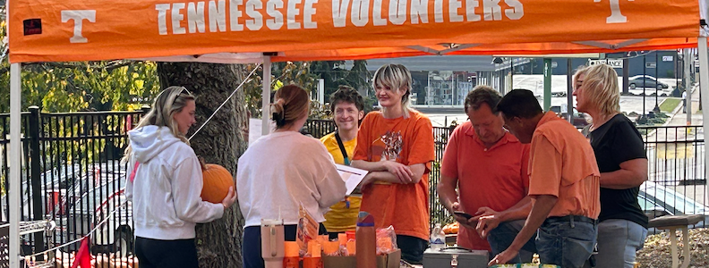 People at a Tennessee Volunteers event stand near an orange tent. Some wear orange, holding items.