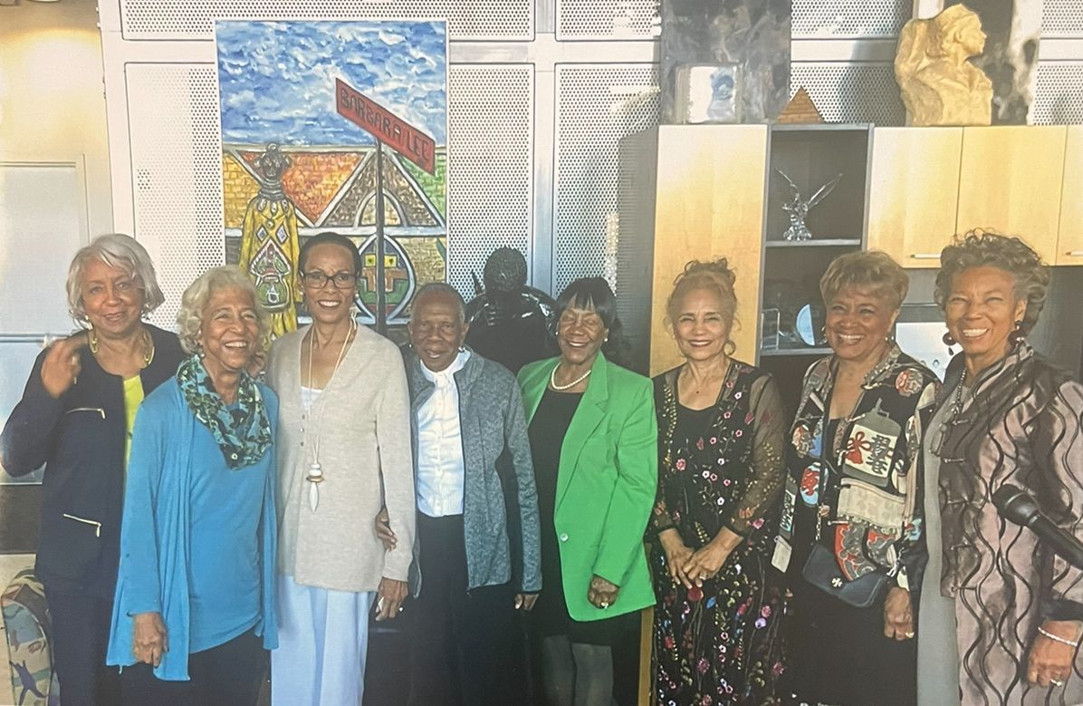 A group of eight people smiling indoors in front of a colorful painting and display cabinets.
