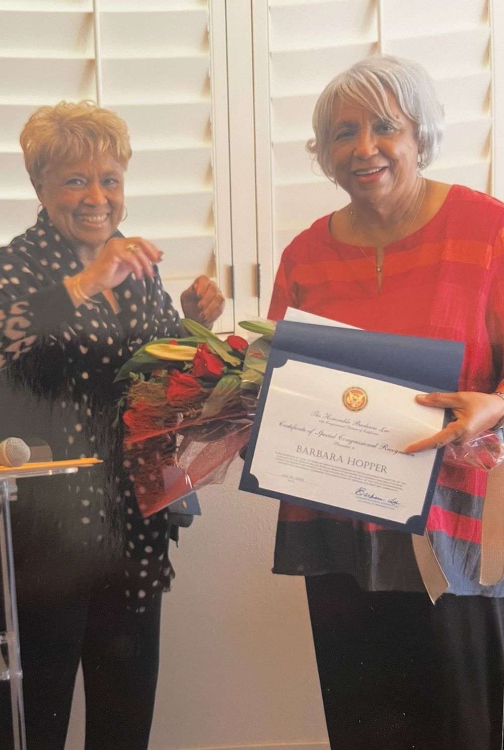 Two women holding flowers and a framed certificate, smiling in front of white shutters.