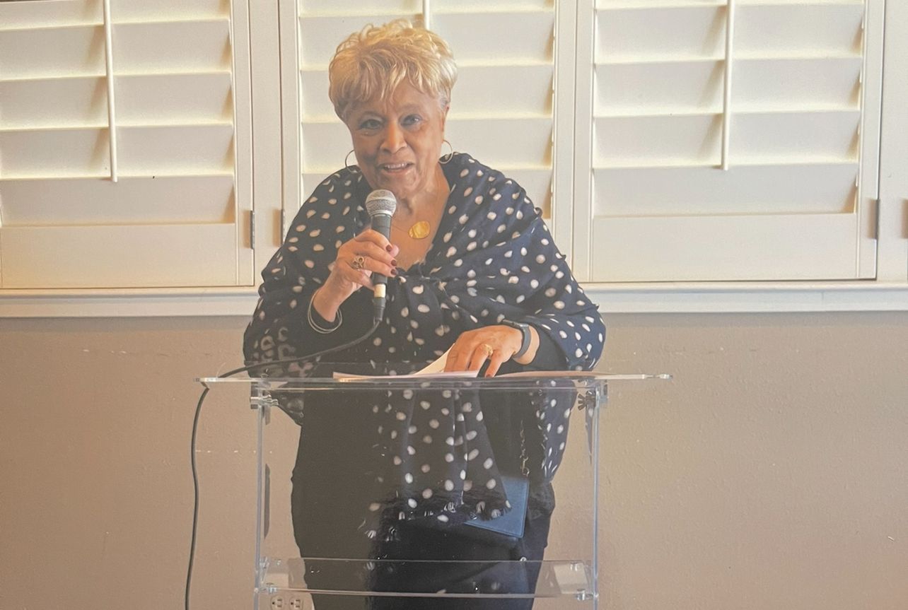Person speaking at a clear podium in front of white shutters, holding a microphone.