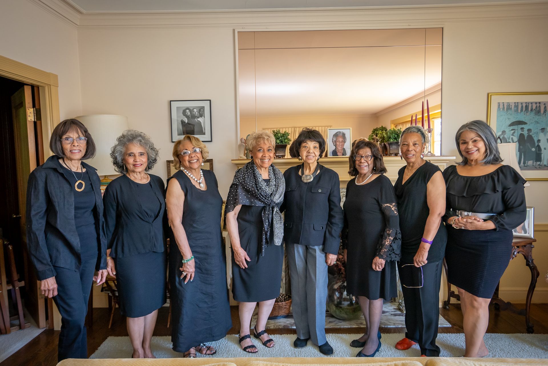 Seven women in black dresses pose together indoors at a formal event.