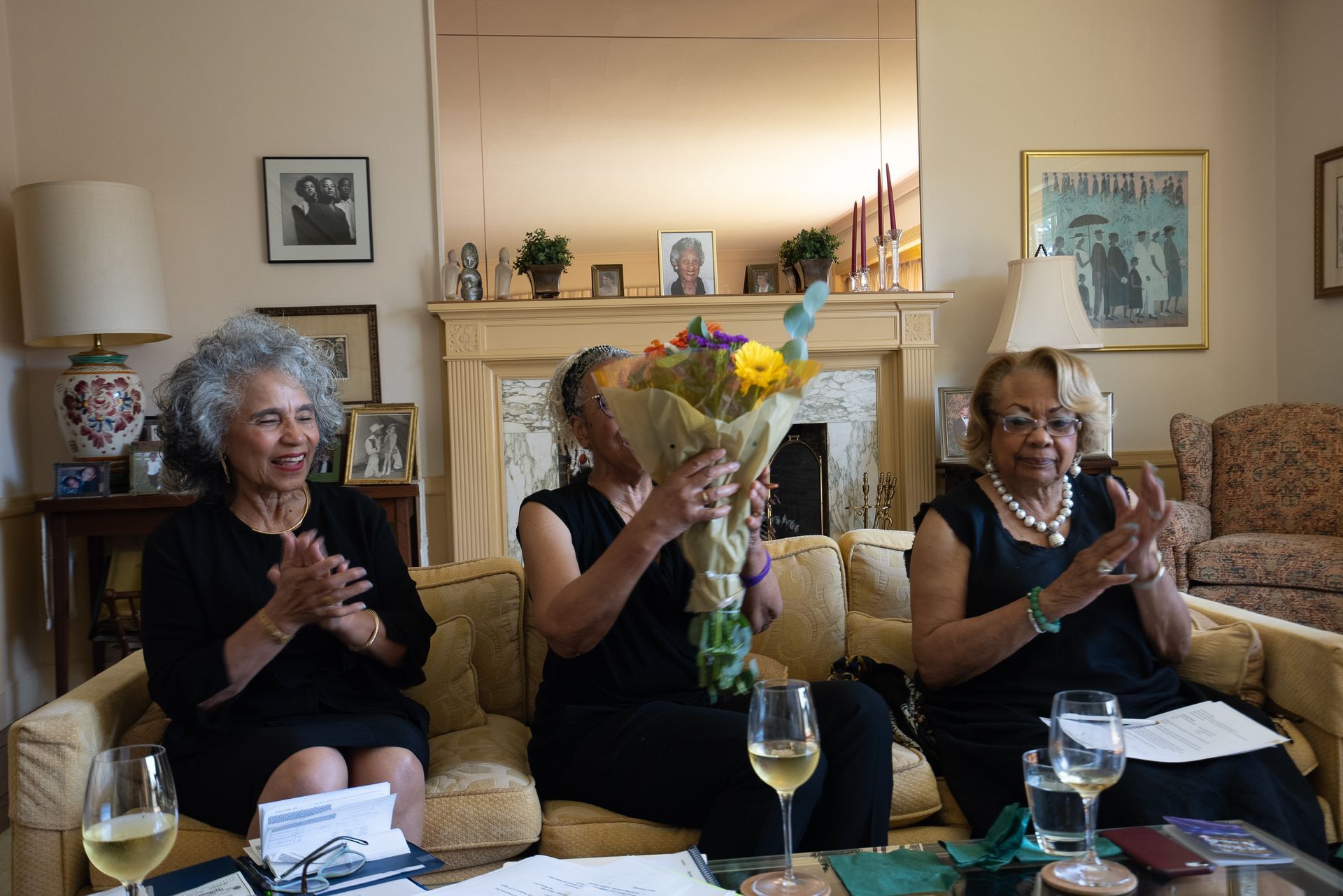 Three women seated on a couch clap around a table with flowers and wine in a cozy living room.