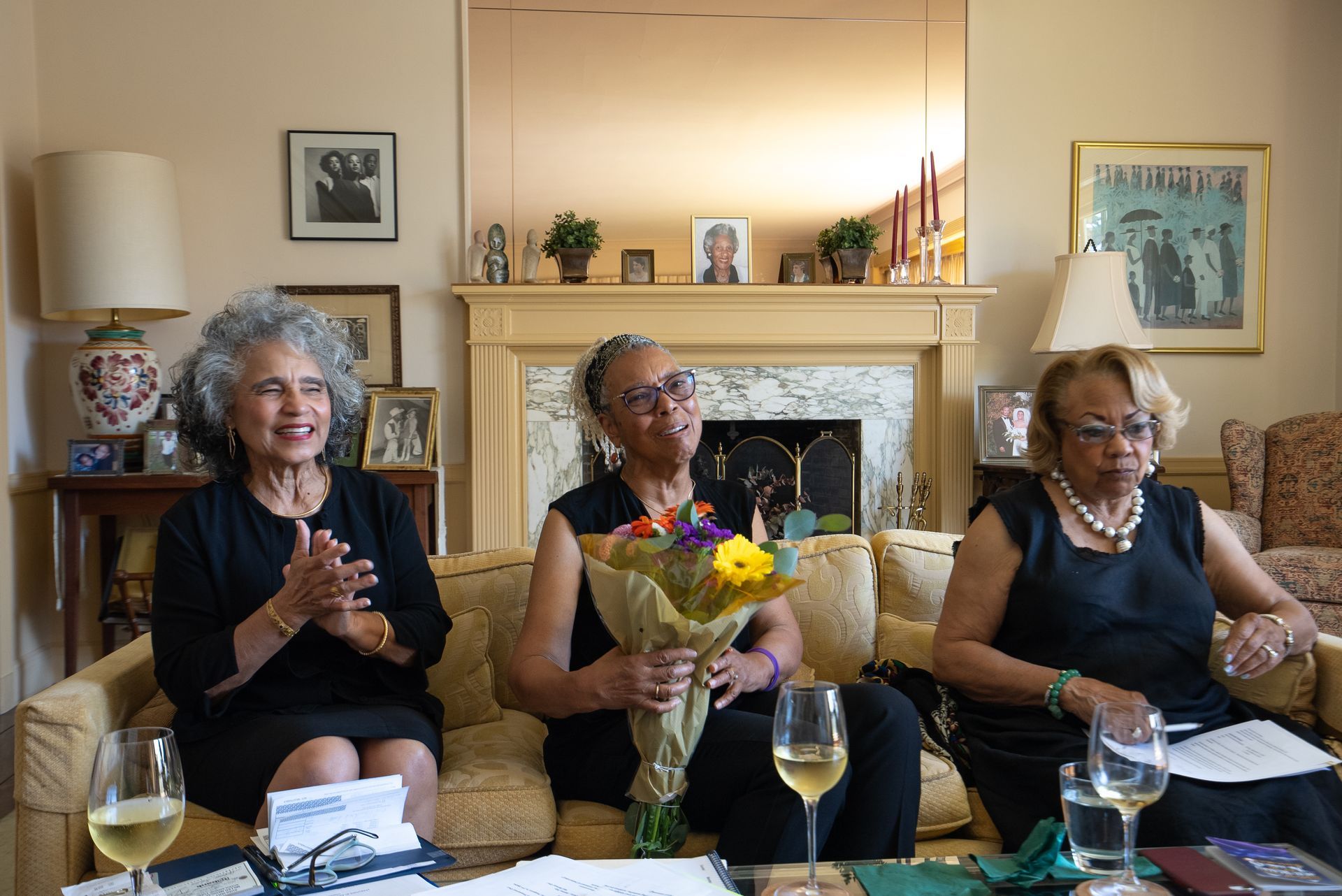 Three women seated in a living room, one holding a bouquet, with drinks and papers on the table.