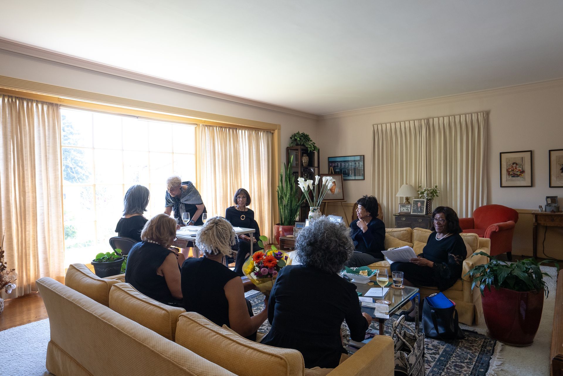 People gathered in a bright living room, seated around a table with flowers and snacks.