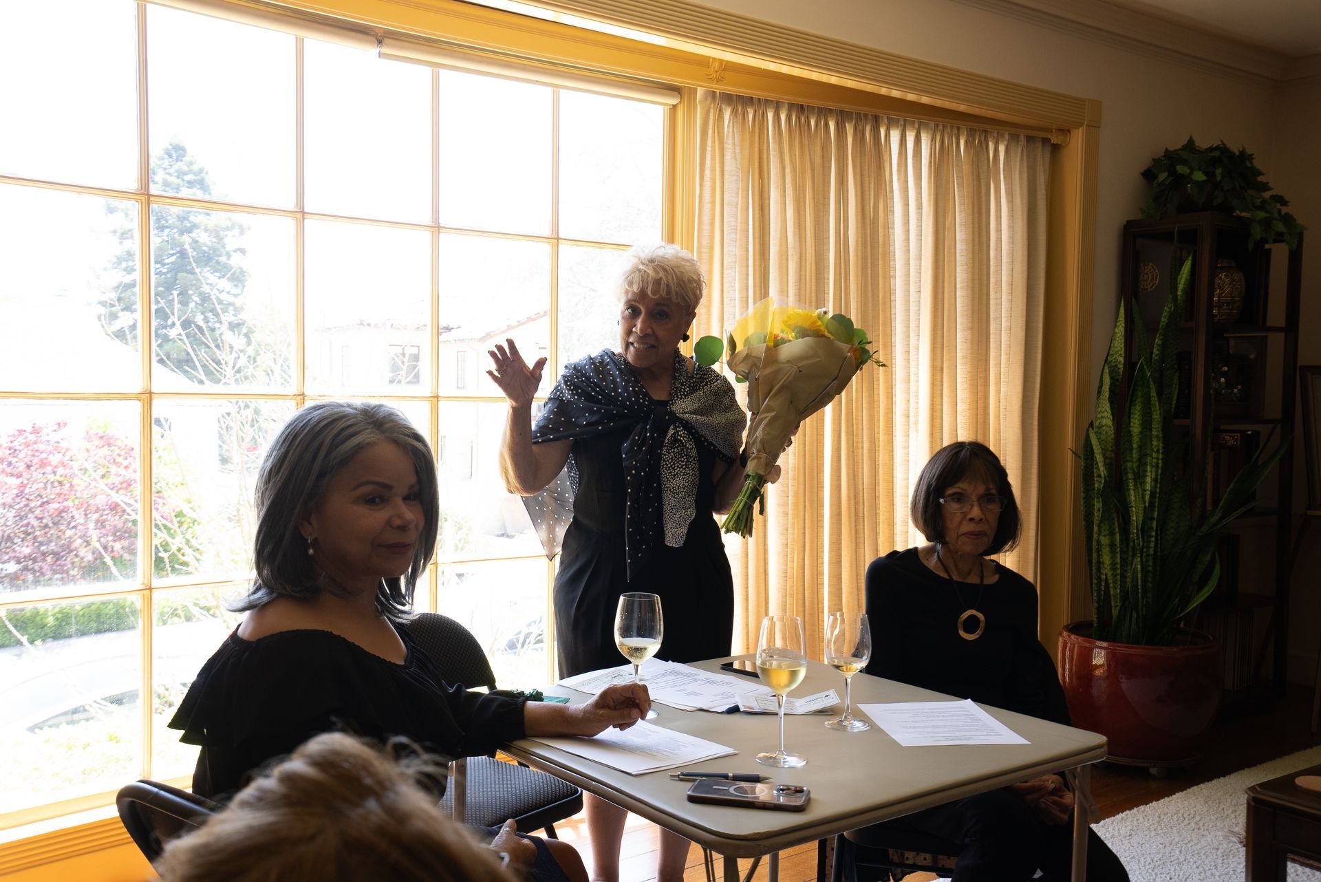 Three people gathered around a table in a sunlit room, one holding flowers near a large window.