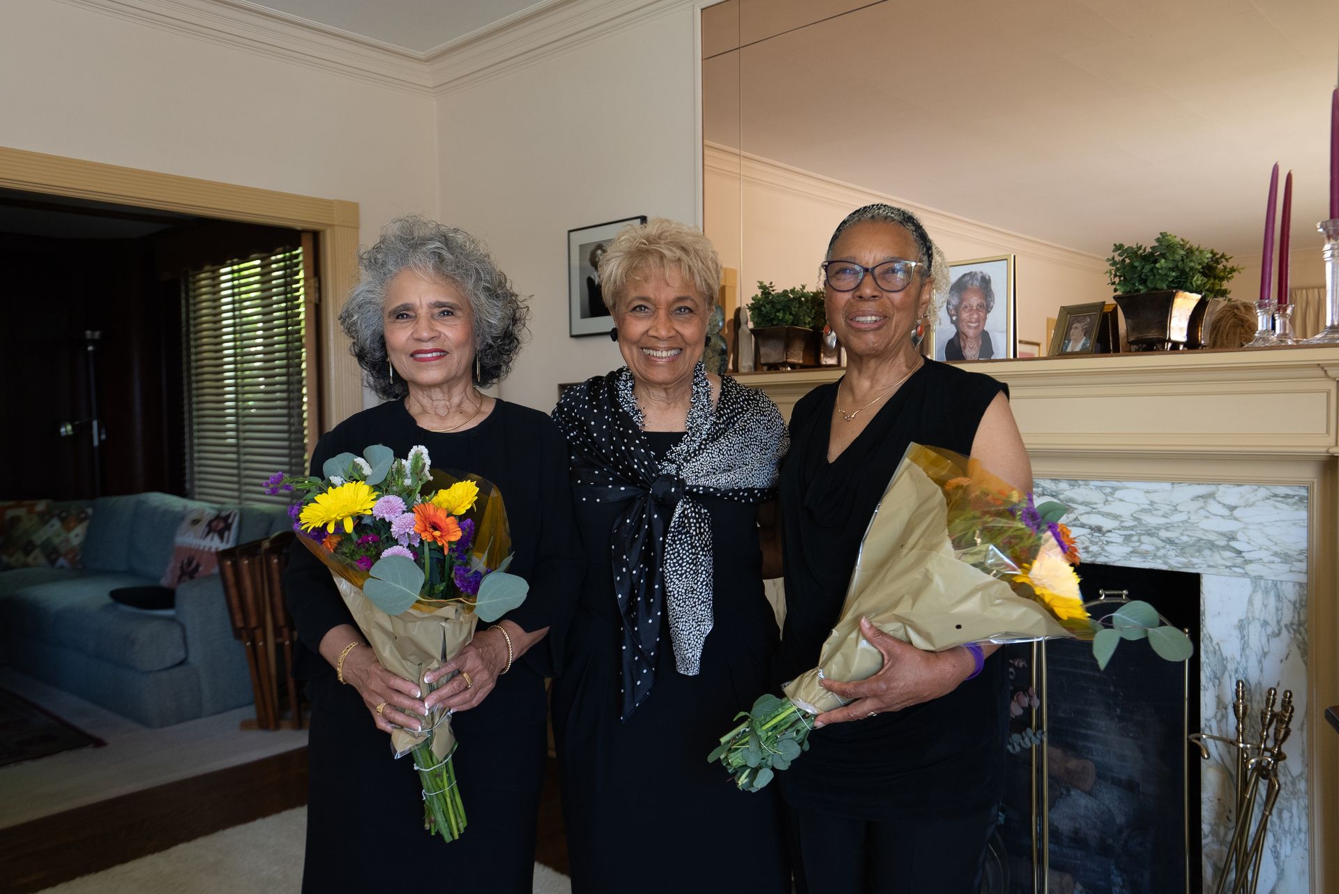Three women in black dresses holding bouquets stand indoors by a fireplace.