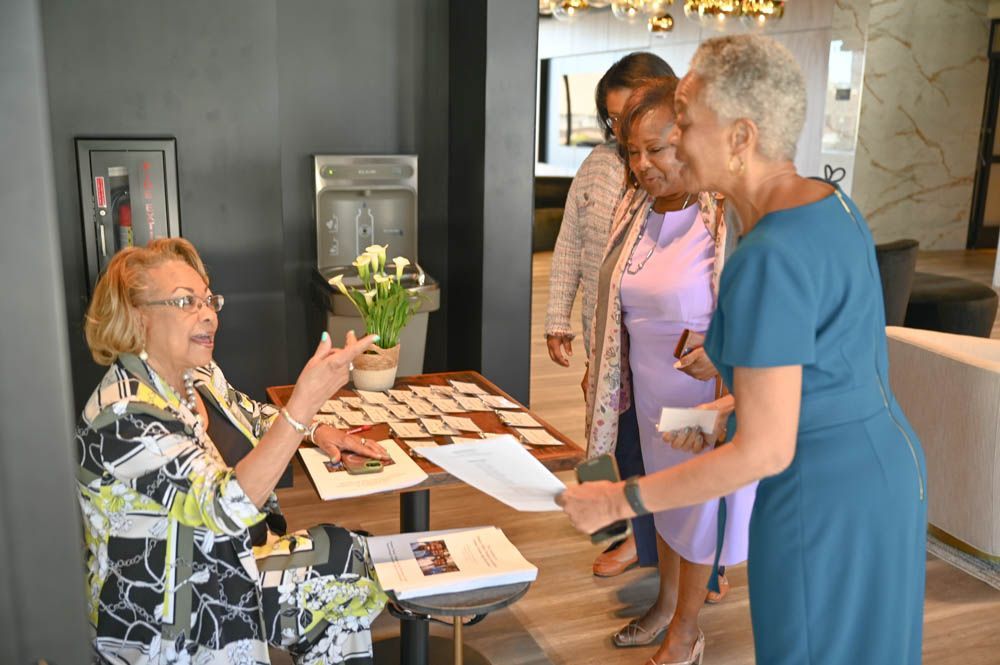 Two women greet a seated volunteer at a registration table in a lobby.