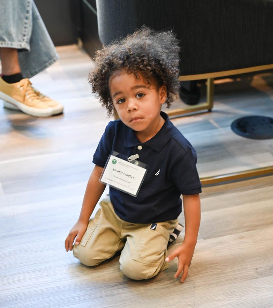 Small child kneeling on a wooden floor in a navy shirt and khaki pants, wearing a name badge.
