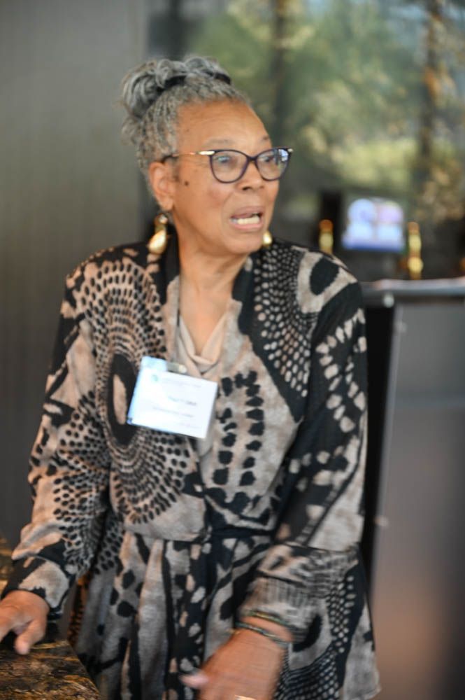 Woman in a patterned black-and-white shawl speaking at an event, wearing glasses and a name badge.