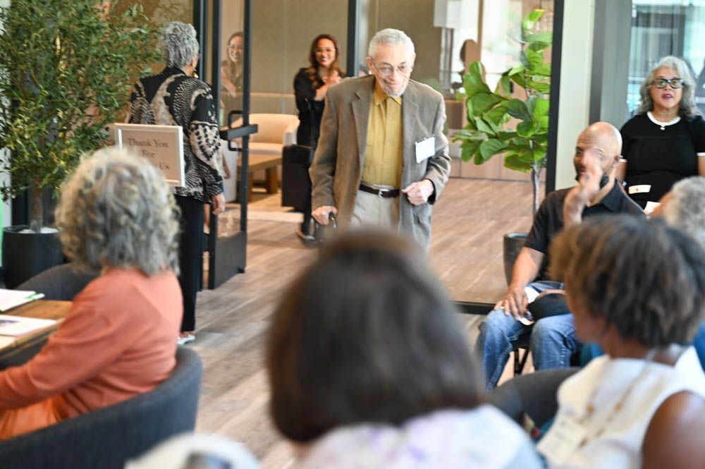 People seated in a meeting room as an older man speaks holding papers, with a woman standing in the background