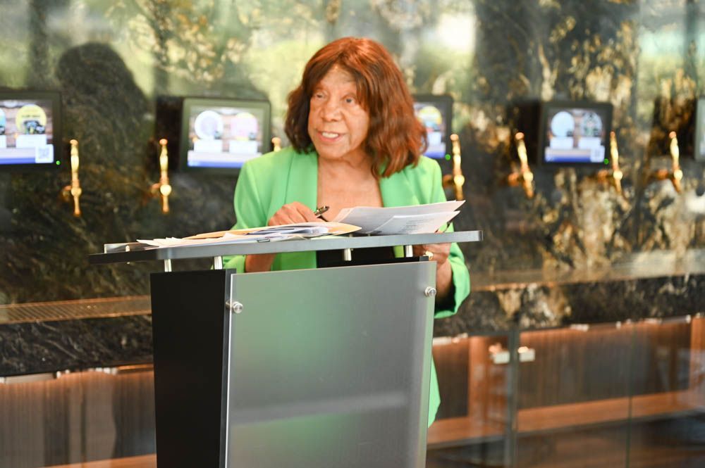 Woman speaking at a podium in a green jacket, with monitors and a marble backdrop behind her.