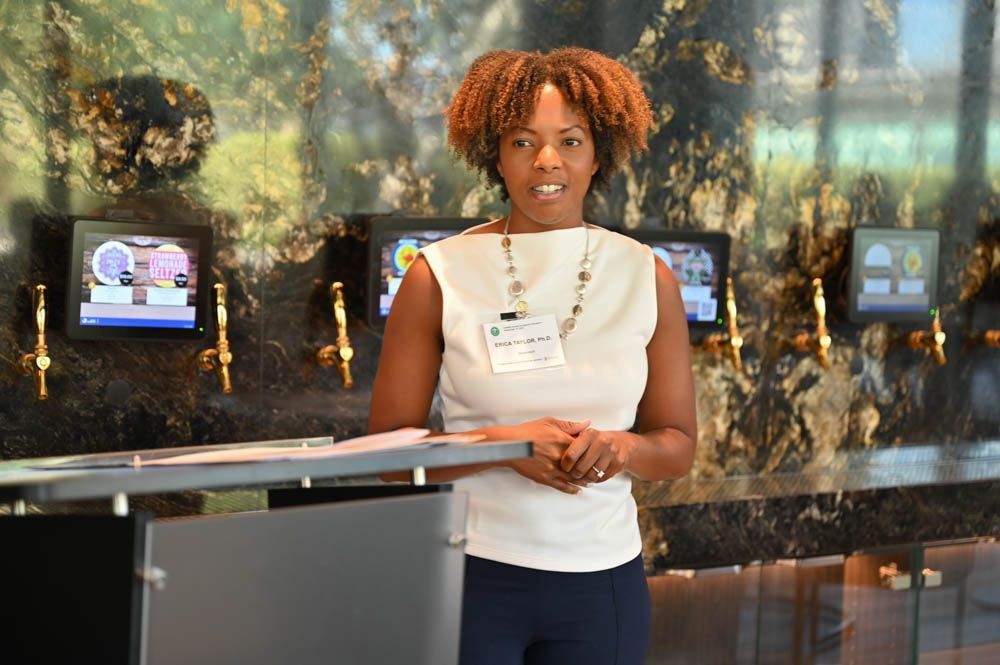 Woman standing at a podium in a lobby, holding papers in front of a wall with small screens.