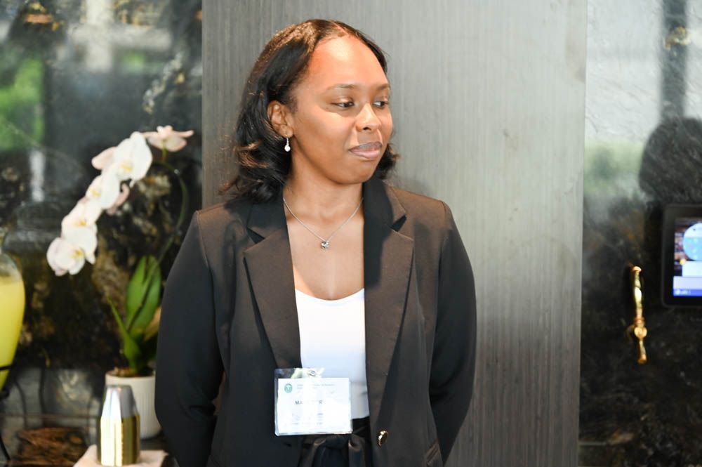 Woman in a black blazer and white top standing indoors with a conference badge and floral arrangement nearby