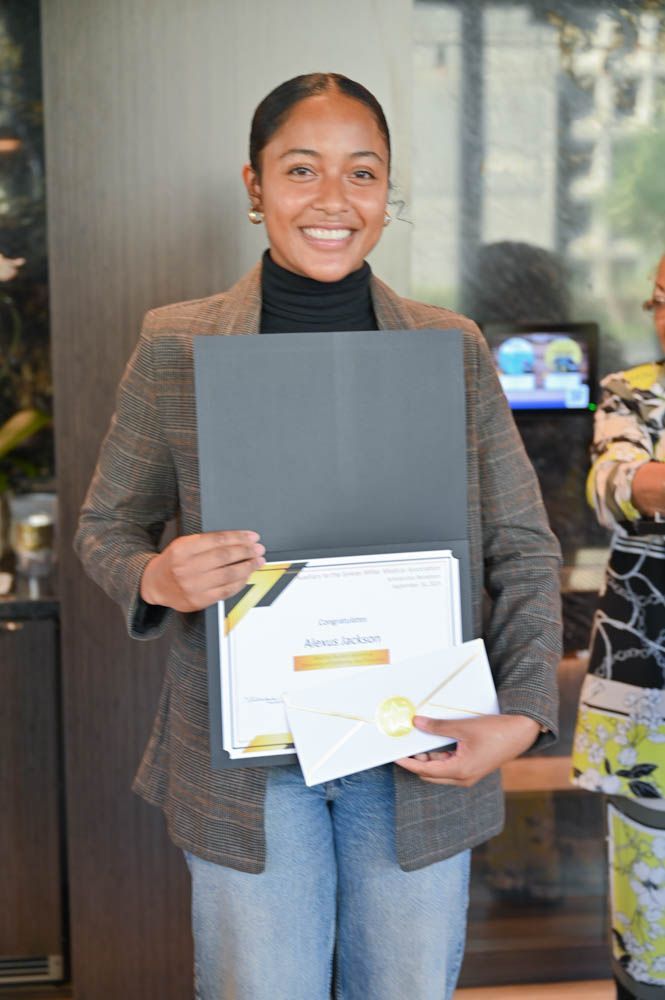 Smiling person holding a certificate folder and envelope indoors, standing by a glass door.