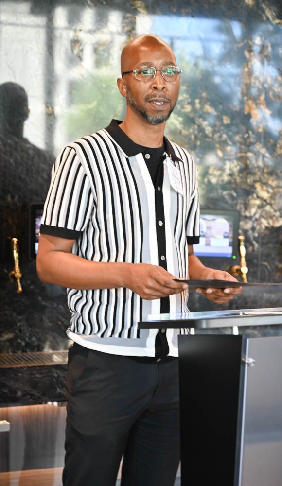 Man standing at a podium, speaking outdoors in a black-and-white striped shirt and glasses.