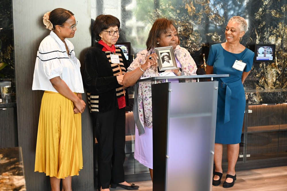 Four women stand at a podium during a speaking event, with a stone wall backdrop.