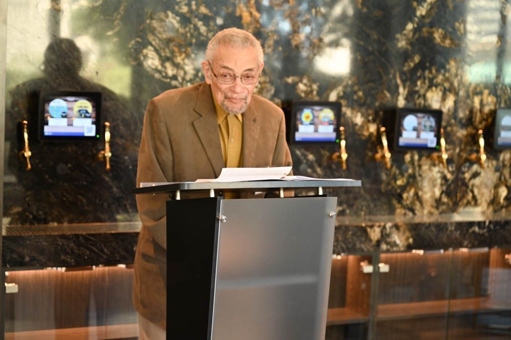 Man speaking at a podium in a dimly lit hall, with small screens behind him.