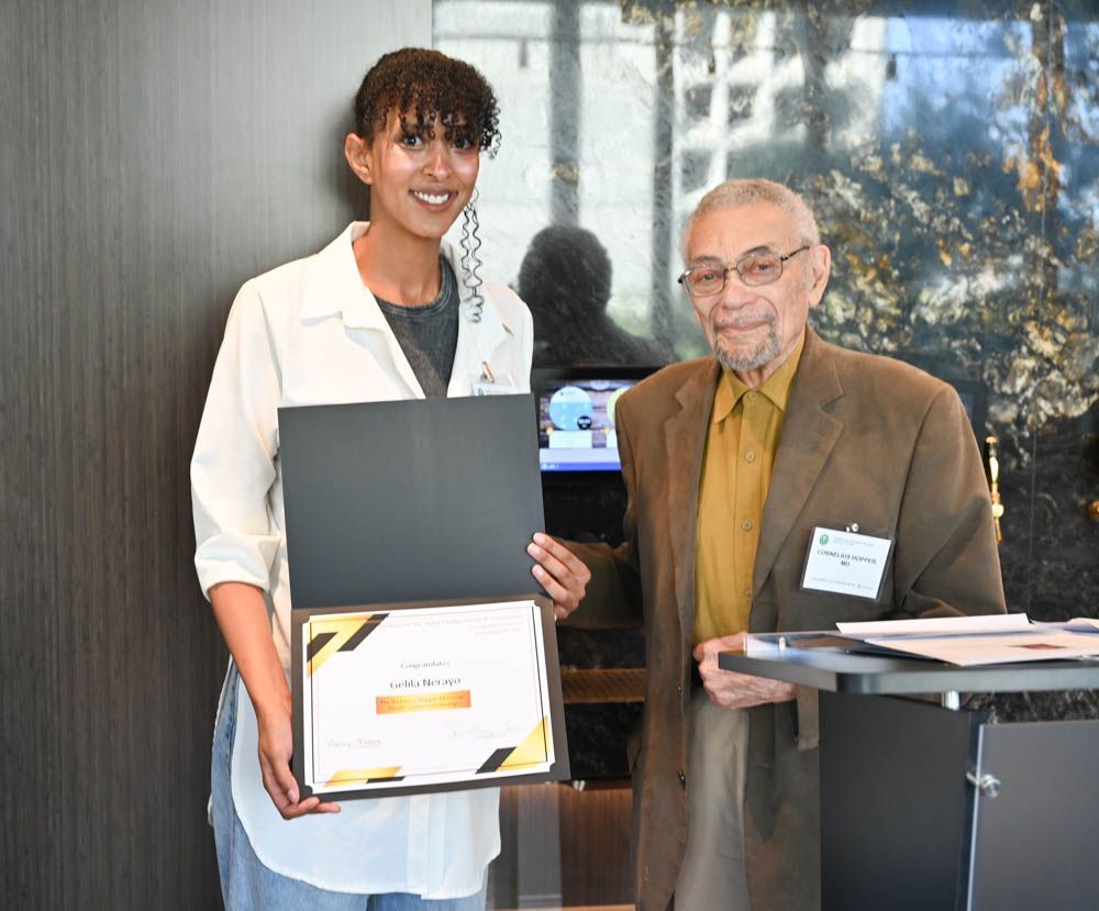 Two people posing indoors, one holding an open certificate folder and the other holding papers.