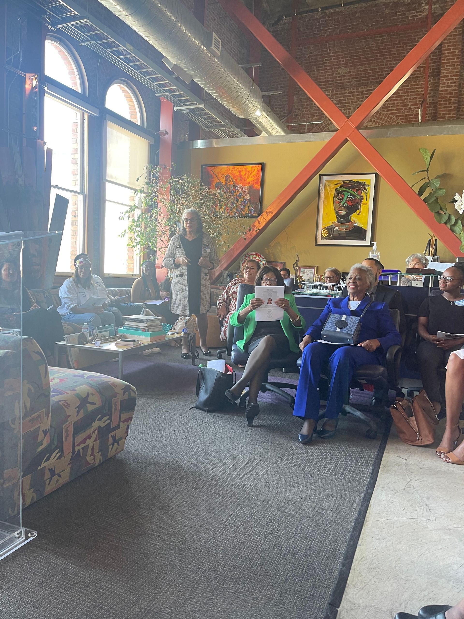 People seated in a sunlit living room with large windows, red X wall decor, and colorful framed art.