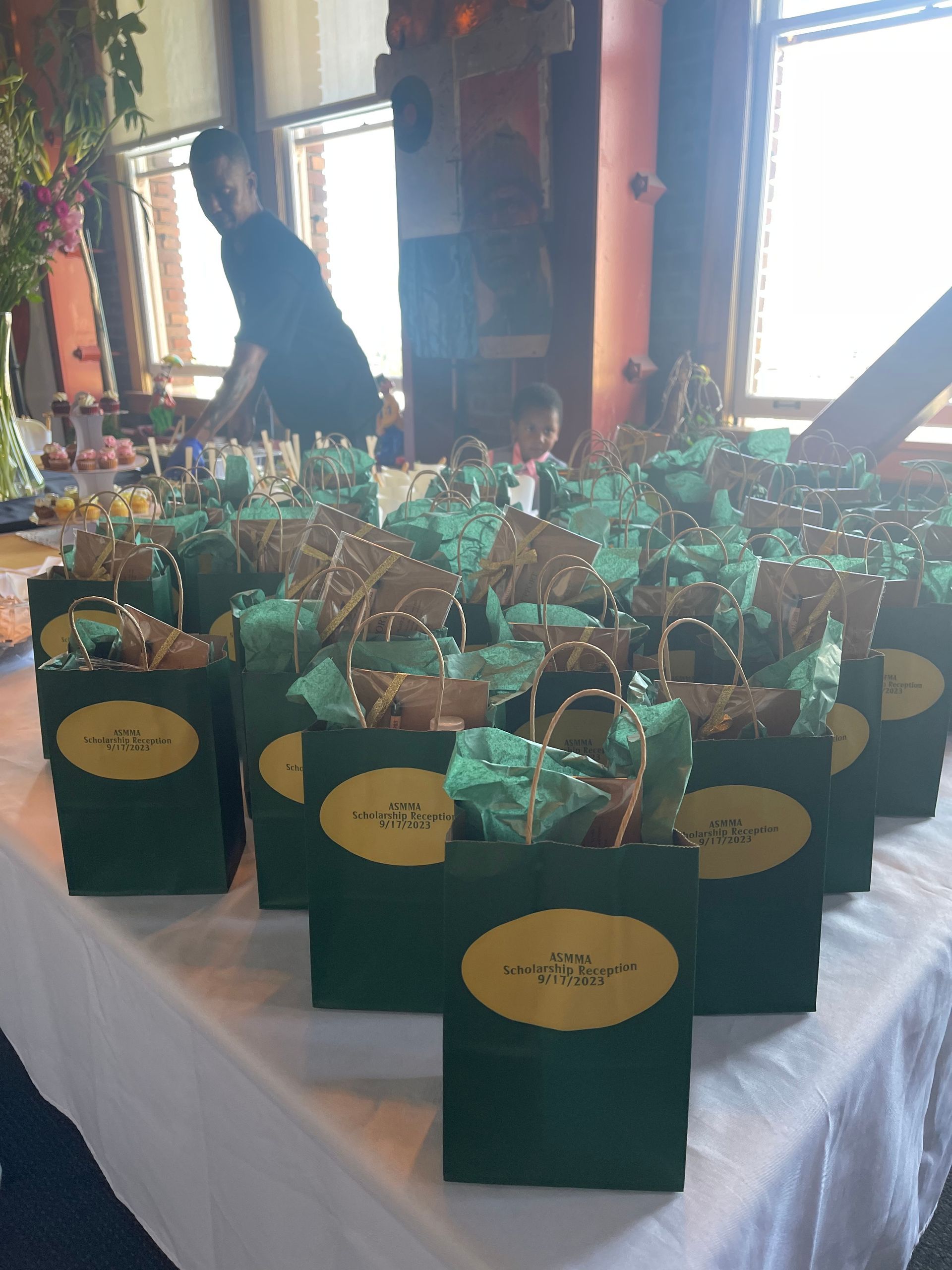 Rows of green gift bags with gold labels on a white table at an indoor event