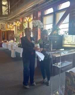Two people standing in a restaurant, holding papers near a table with white tablecloths and flowers at Auxiliary to the Sinkler Miller Medical Association
