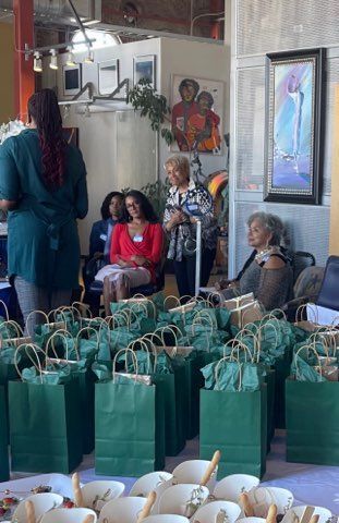 Gift bags lined up on a table in a gathering space, with several people seated and standing nearby.