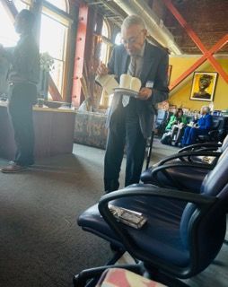 Man in a suit carrying a tray in a busy airport waiting area, with empty blue seats in foreground