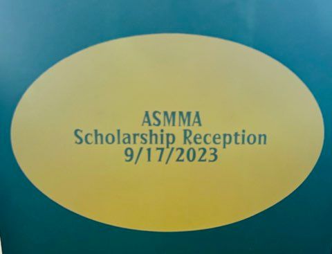 Yellow oval sign reading “ASMMA Scholarship Reception 9/17/2023” on a teal background
